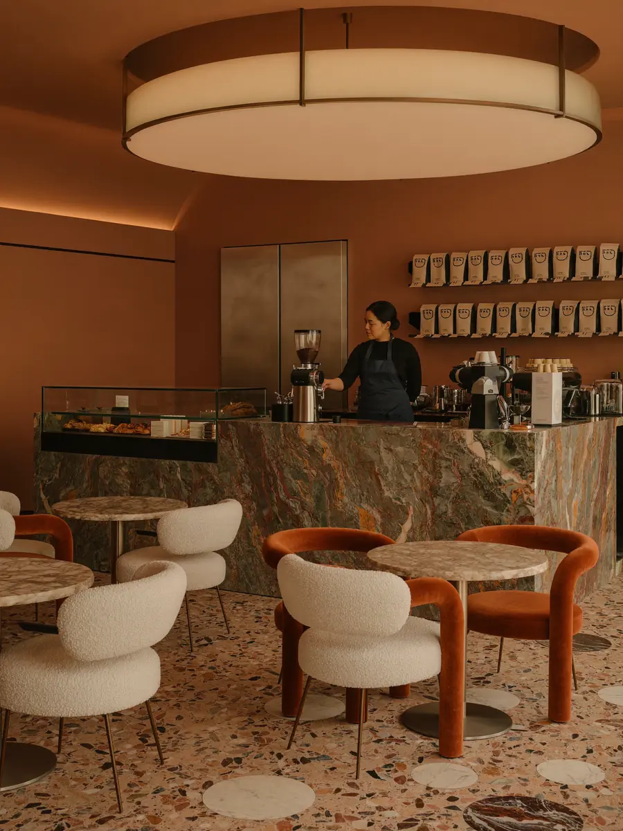 Barista preparando café en una barra de mármol con vitrina de pastelería y bolsas de café en la estantería